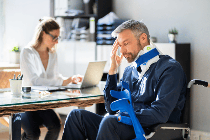 Man in a wheelchair with a neck brace holds a crutch in an office setting, while a car accident lawyer myrtle beach works on a laptop in the background.